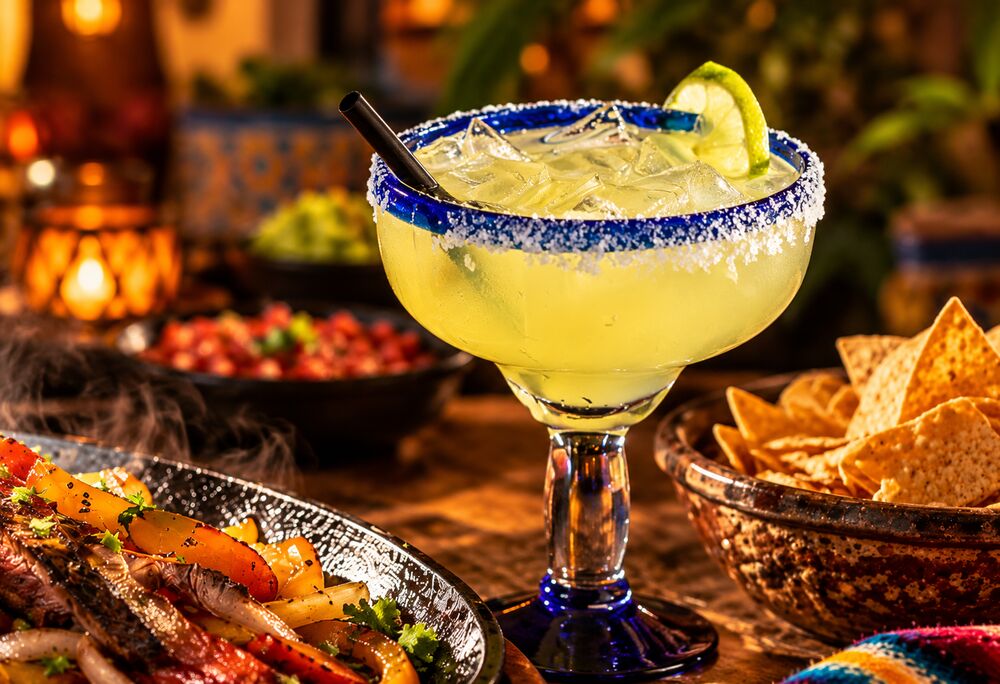 Salt-rimmed margarita in a blue-rimmed glass with lime, next to chips, salsa and sizzling fajitas